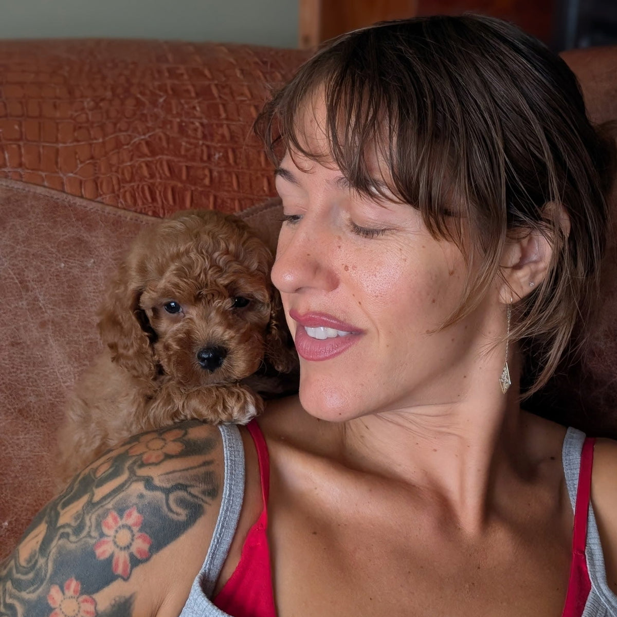A woman with short brown hair and a floral tattoo on her arm rests against a brown couch, smiling next to a small curly-haired brown dog. She is wearing a gray tank top with a red lining.