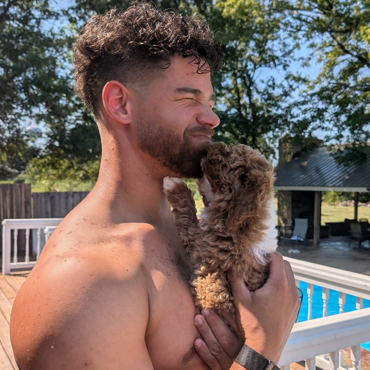 A shirtless man holding a small curly-haired brown and white dog on a wooden deck next to a swimming pool, with trees and a house in the background.