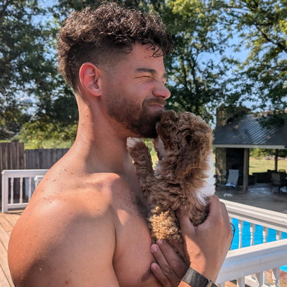 A shirtless man holding a small curly-haired brown and white dog on a wooden deck next to a swimming pool, with trees and a house in the background.