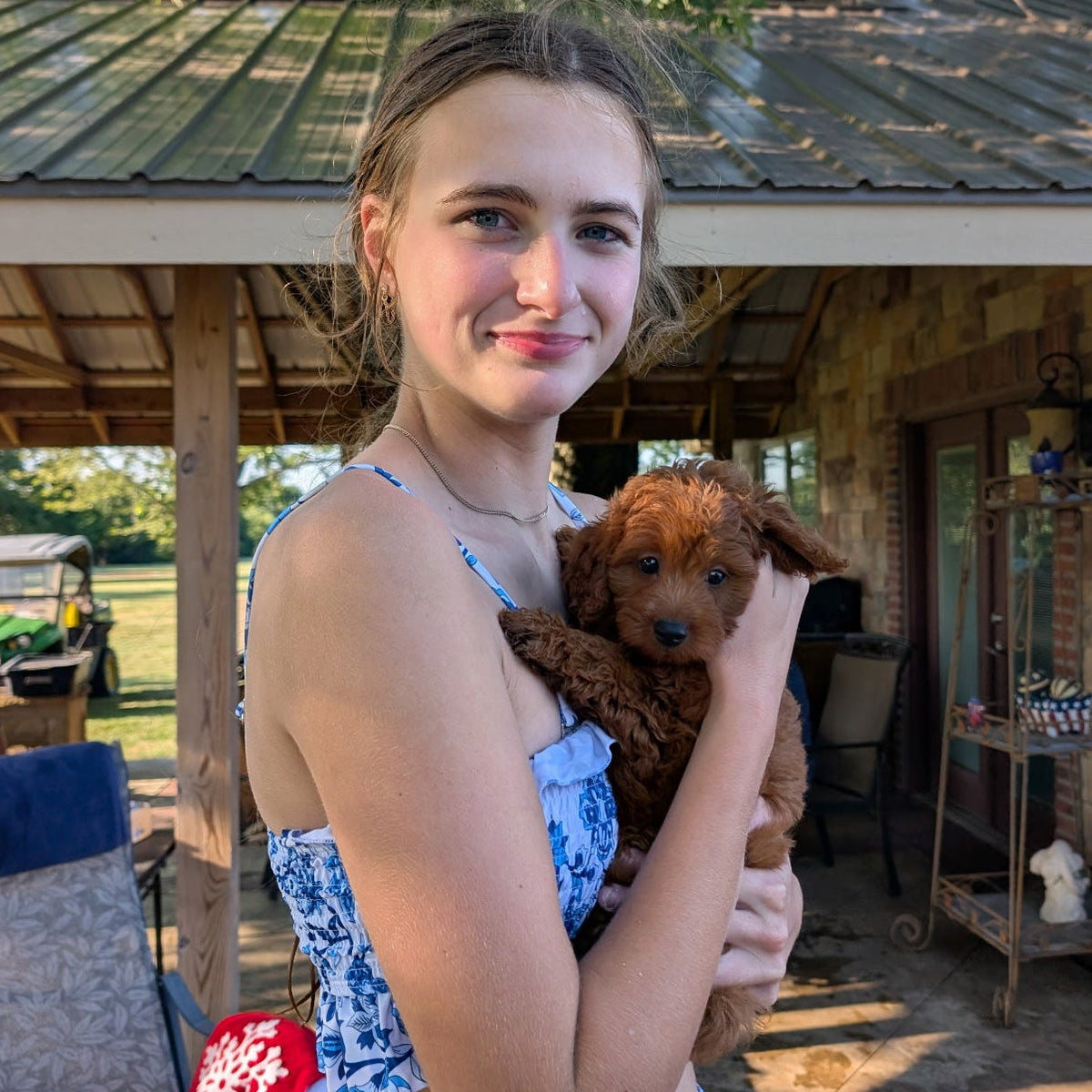 A woman holding a small brown puppy on a rustic outdoor porch. She is wearing a blue and white floral print crop top and matching skirt.