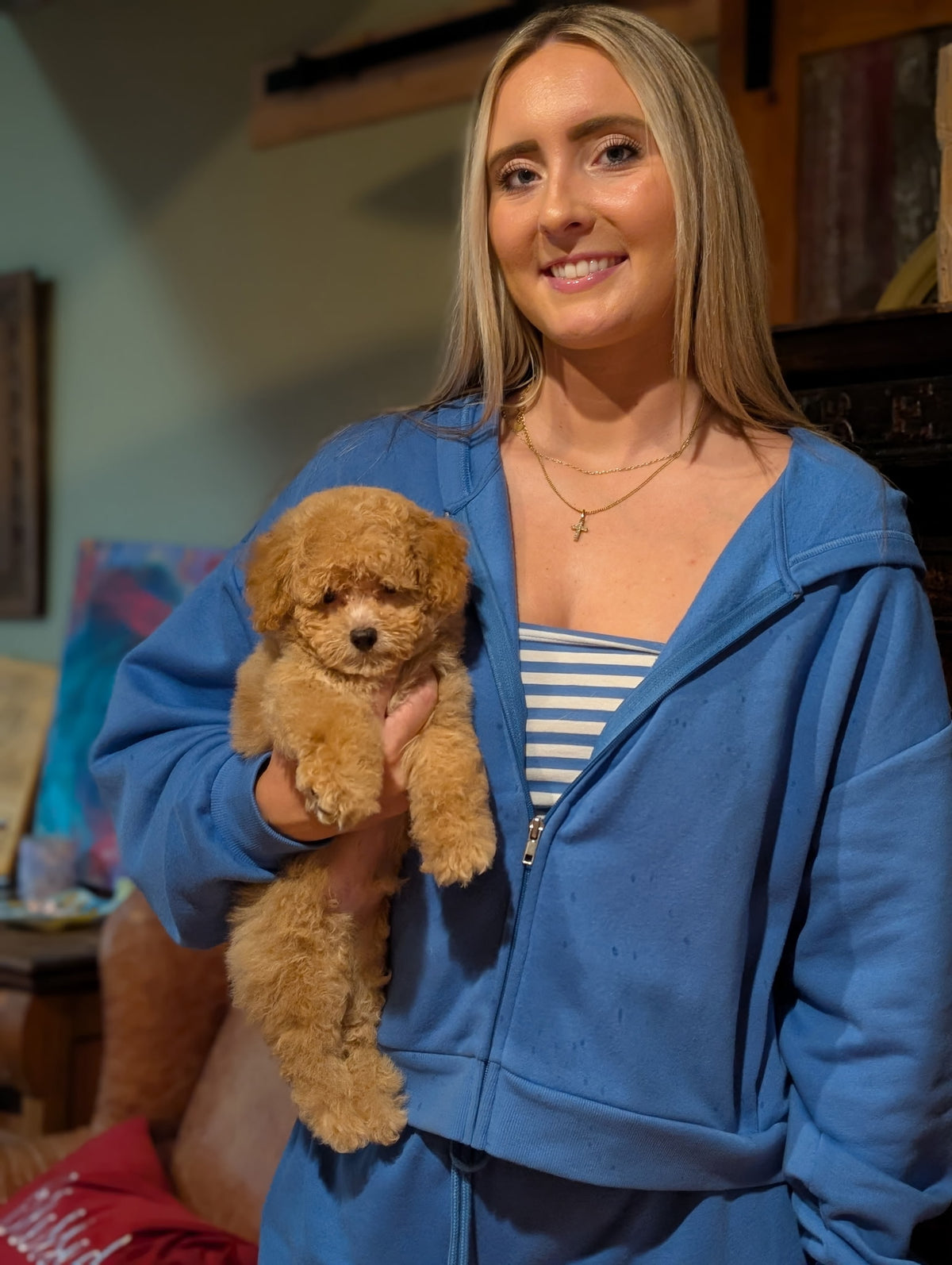 A woman with blonde hair wearing a blue zip-up hoodie and a striped top, holding a small fluffy golden-brown puppy in an indoor setting.